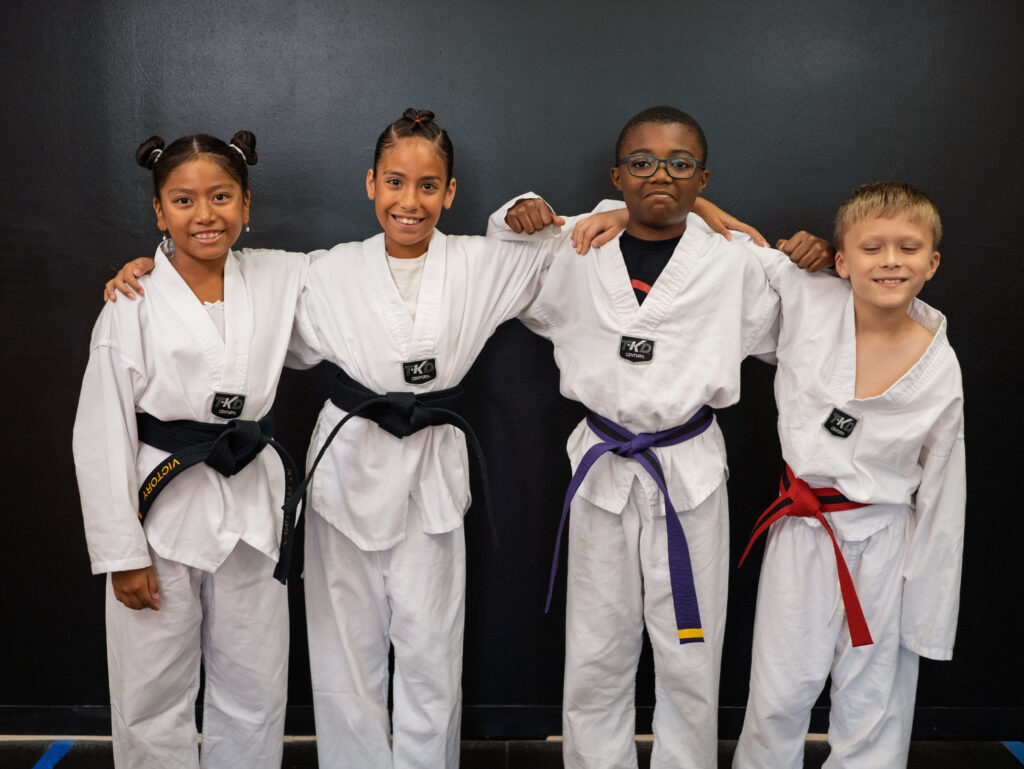 Four smiling children in white martial arts uniforms stand side by side with their arms around each other against a dark studio background. They wear different belt colors, including black, purple, and red.