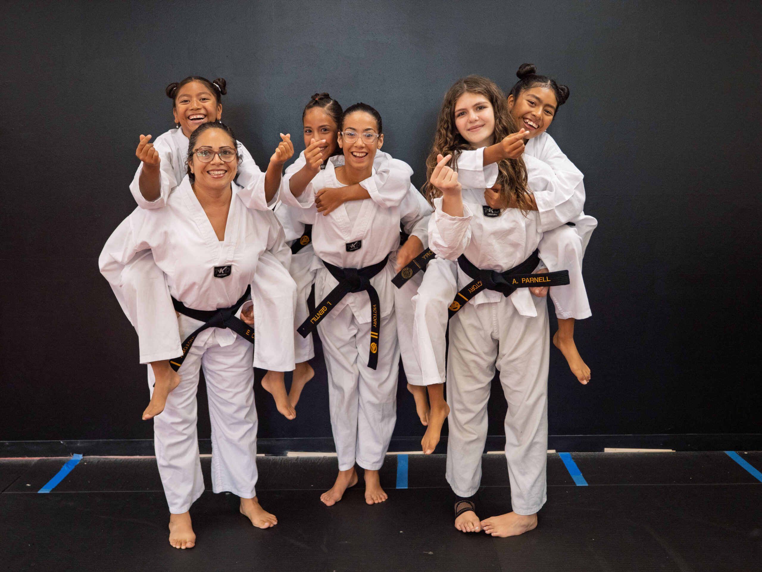 Group photo of three martial arts instructors in white uniforms and black belts, each carrying one or two smiling children in matching uniforms against a dark studio wall. The image is framed with a blue border and a blue check mark icon in the top right corner, as shown on a page with other party-themed photos above and below.