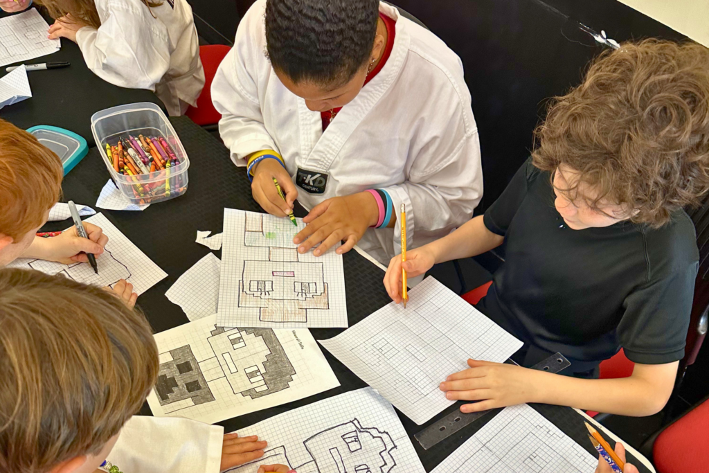 Photo of a MacBook Pro displaying an image of children gathered around a table doing art activities. The kids are drawing on graph paper with pencils and crayons, with several block-style designs spread across the table. The laptop sits indoors in front of a softly blurred background with a grand piano and bright windows.