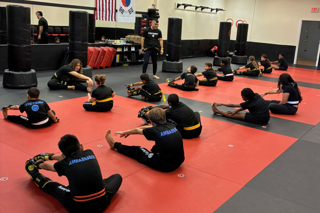 Photo of a MacBook Pro displaying a martial arts camp image in Preview. On the laptop screen, an instructor leads a group of children in black uniforms stretching on red-and-black mats inside a martial arts studio with punching bags and flags on the wall. The laptop is sitting indoors in front of a softly blurred background with a grand piano and bright windows.